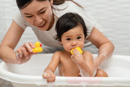 mother bathing her cheerful infant baby with foam bubbles and playing rubber duck in a bathtubの写真素材