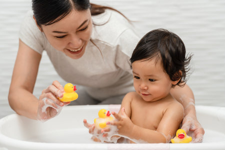 mother bathing her cheerful infant baby with foam bubbles and playing rubber duck in a bathtubの写真素材