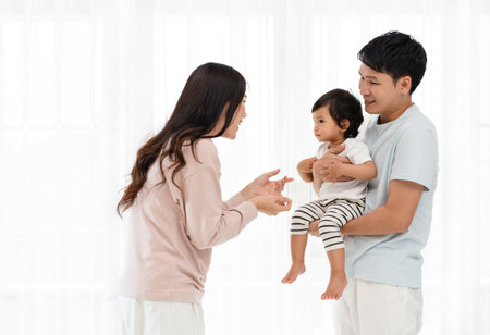 cheerful father and mother holding and playing with toddler baby on white window background. happy family conceptの写真素材