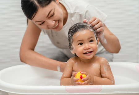 mother bathing and washing her infant baby hair with shampoo while playing a rubber duck toy in a bathtubの写真素材