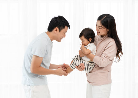 cheerful father and mother holding and playing with toddler baby on white window background. happy family conceptの写真素材