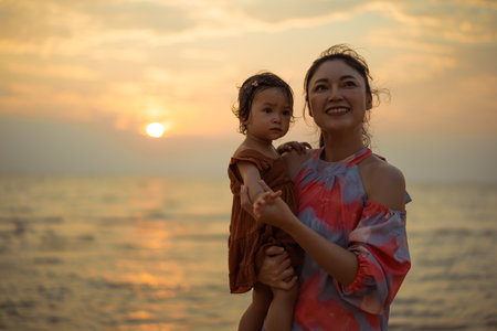 mother holding and talking toddler baby girl on the sea beach at sunsetの写真素材