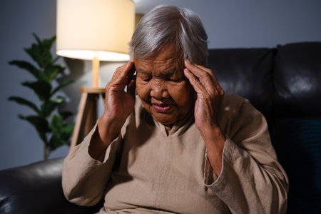 senior woman having a headache pain while sitting on a sofa in living room at nightの写真素材