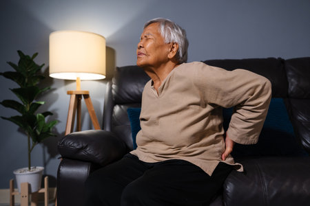 senior woman suffering from backache while sitting on a sofa in living room at nightの写真素材