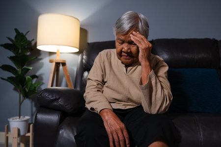 senior woman having a headache pain while sitting on a sofa in living room at nightの写真素材