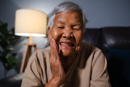 senior woman feels strong toothache while sitting on a sofa in the living room at nightの写真素材