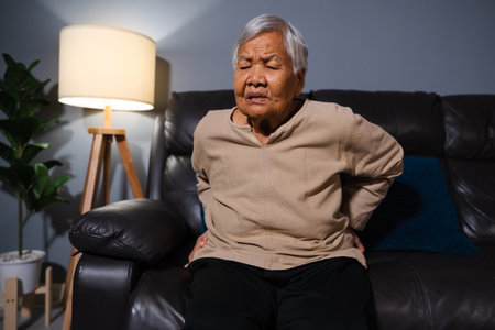 senior woman suffering from backache while sitting on a sofa in living room at nightの写真素材