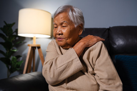 senior woman suffering from neck and shoulder pain while sitting on a sofa in living room at nightの写真素材