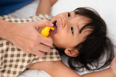 mother or doctor spraying medicine in mouth of toddler girl. baby sore throat treatment conceptの写真素材