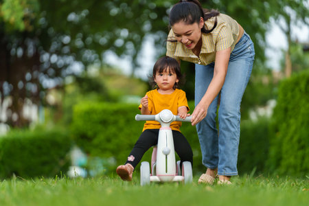 mother teaches her baby toddler girl rides a plastic motorcycle toy in the parkの写真素材