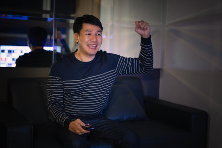 cheerful man watching sport TV with arm raised on a sofa at nightの写真素材