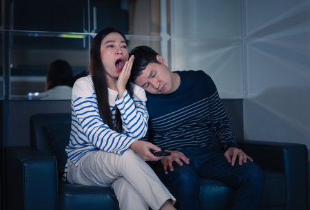 sleepy couple yawning and sleeping while watching TV movie on a sofa at nightの写真素材