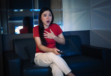 shocked and scared woman is watching horror movie TV on a sofa at nightの写真素材