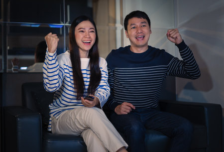 cheerful couple watching sport TV with arm raised on a sofa at nightの写真素材