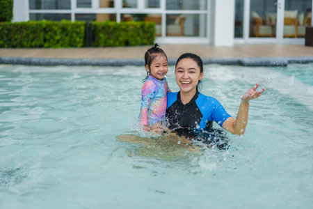 happy mother holding toddler baby girl in her arms while playing water splashing in a swimming poolの写真素材