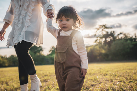 little toddler baby girl holding her mother's hand and walking in a grass field at sunsetの写真素材