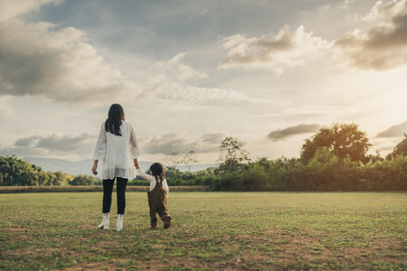 happy mother walking with her toddler baby girl on a grass field at sunsetの写真素材