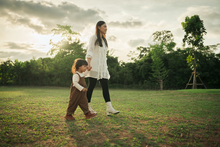 happy mother walking with her toddler baby girl on a grass field at sunsetの写真素材