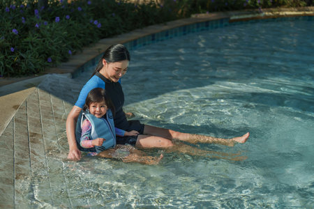 happy mother with toddler baby girl in life jacket sitting in edge of the swimming poolの写真素材