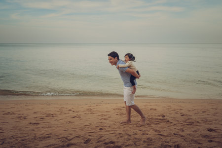 father giving piggyback ride to toddler baby girl walking on the sea beach in Pattaya, Thailandの写真素材