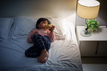 toddler baby girl drinking milk from bottle on a bed at nightの写真素材