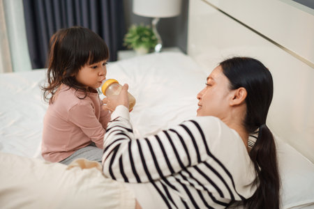 mother feeding baby girl with milk in bottle on a bedの写真素材