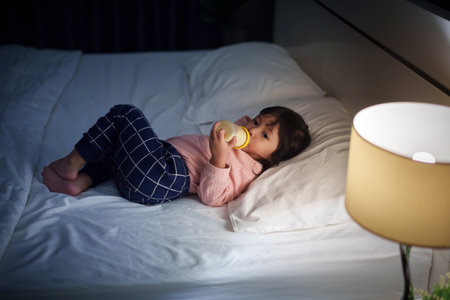 toddler baby girl drinking milk from bottle on a bed at nightの写真素材