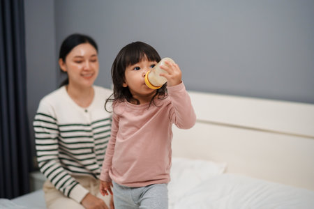 toddler girl drinking milk from bottle with her mother on a bedの写真素材