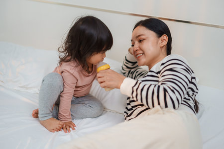 mother feeding baby girl with milk in bottle on a bedの写真素材