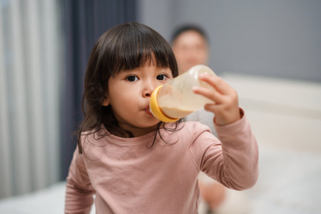 toddler girl drinking milk from bottle with her mother on a bedの写真素材