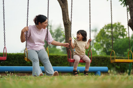cheerful toddler girl with mother playing on a swing at outdoor playgroundの写真素材