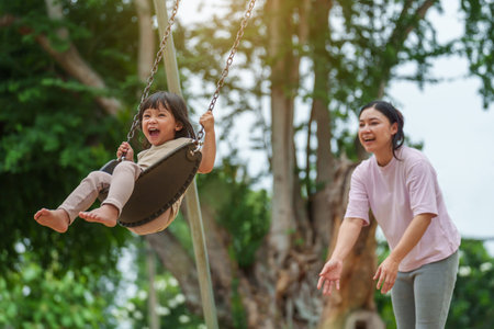 cheerful toddler girl with mother playing on a swing at outdoor playgroundの写真素材