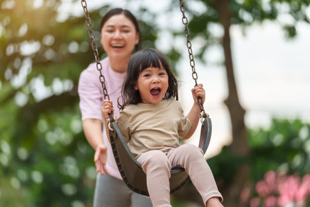 cheerful toddler girl with mother playing on a swing at outdoor playgroundの写真素材