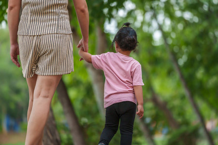toddler girl walking and holding hand with her mother on sidewalk at parkの写真素材