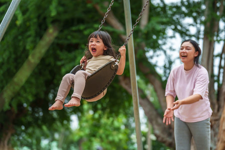 cheerful toddler girl with mother playing on a swing at outdoor playgroundの写真素材