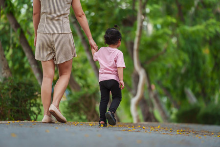 toddler girl walking and holding hand with her mother on sidewalk at parkの写真素材