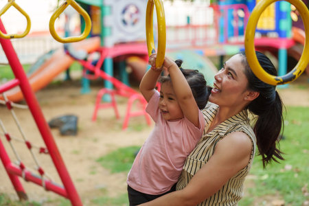 mother holding her daughter to hanging on playground rings in the parkの写真素材