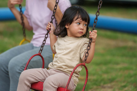 cheerful toddler girl with mother playing on a swing at outdoor playgroundの写真素材