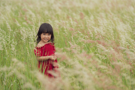 cheerful toddler girl in red dress playing grass flower in fieldの写真素材