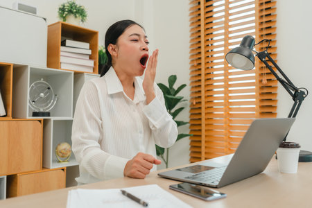 sleepy young woman yawning while working with laptop computer at home officeの写真素材