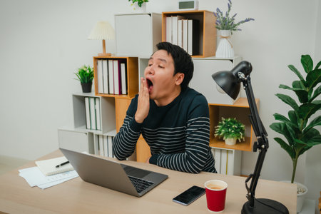 sleepy young man yawning while working with laptop computer at home office