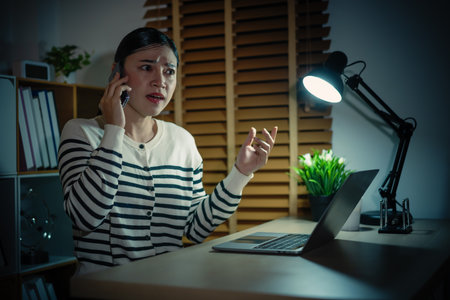 stressed woman talking with smartphone while working with laptop at a nightの写真素材