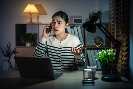 stressed woman talking with smartphone while working with laptop at a nightの写真素材