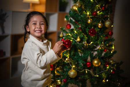 happy toddler girl decorating toy ball on Christmas tree in home at nightの写真素材
