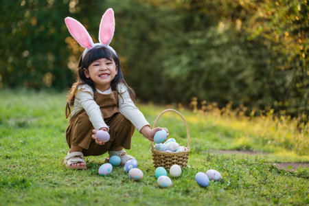 happy child girl wearing bunny ears playing and hunting for Easter eggs into a basket on green grass...