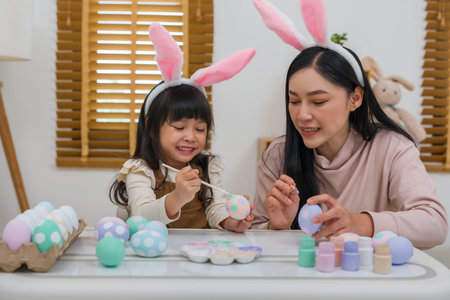mother and preschool child girl wearing bunny ears painting and decorating egg  together at their...