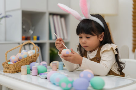 preschool child girl wearing bunny ears painting and decorating colorful egg at the home on easter...