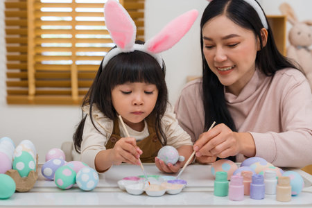 mother and preschool child girl wearing bunny ears painting and decorating egg  together at their...