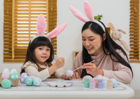 mother and preschool child girl wearing bunny ears painting and decorating egg  together at their...
