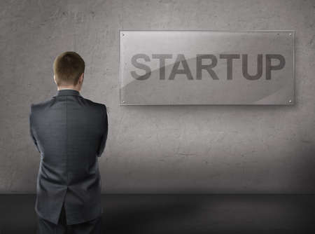 Business man standing front of the wall with large glass plate, word STARUP on it.の写真素材